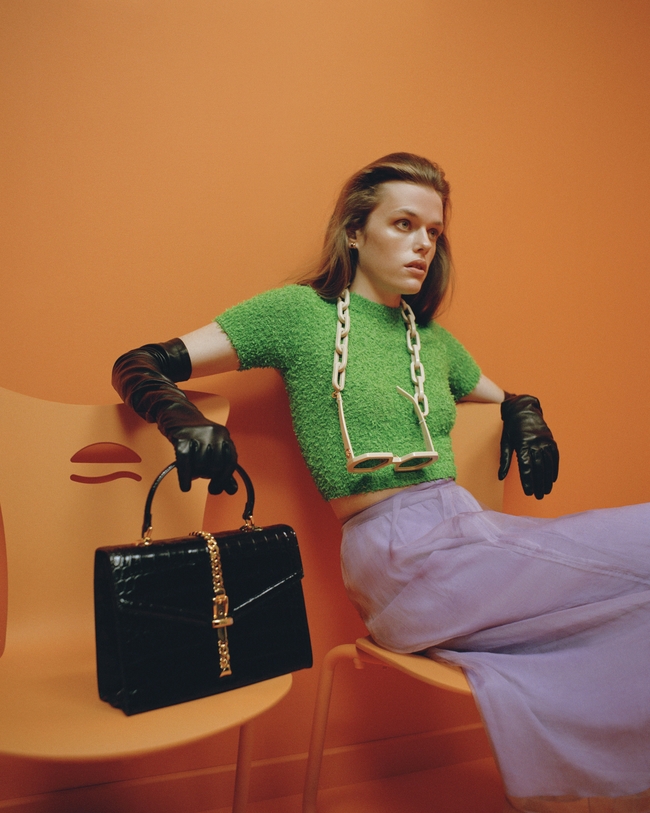 حقيبة يد جلديّة Sylvie 1969 Top Handle Bag  وبلوزة من مزيج القطن وقفّازات جلديّة ونظّارات بسلاسل من الأسيتات وتنوّرة حريريّة، كلّها من Gucci. وأقراط نحاسيّة من Louis Vuitton